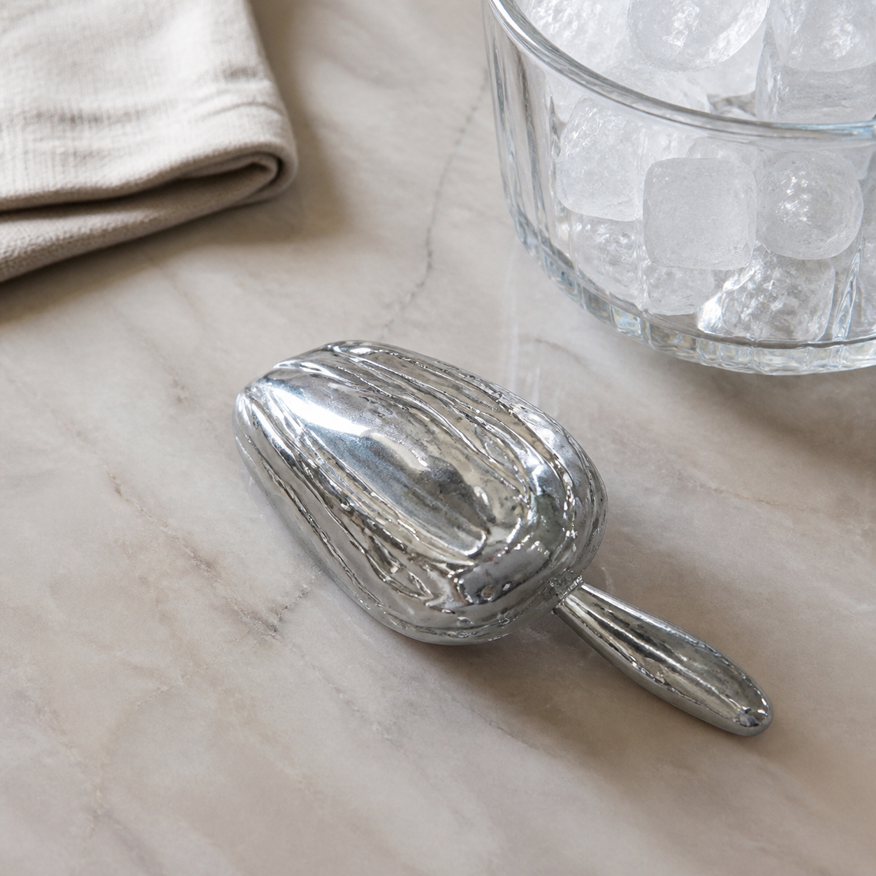 Silver metal ice scoop lying bowl-down on a marble counter beside a glass bucket of ice