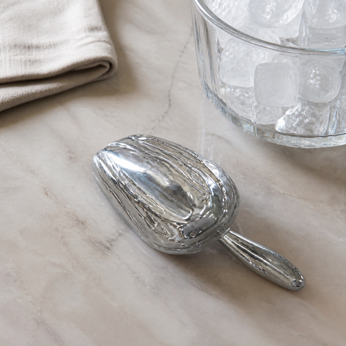 Silver metal ice scoop lying bowl-down on a marble counter beside a glass bucket of ice