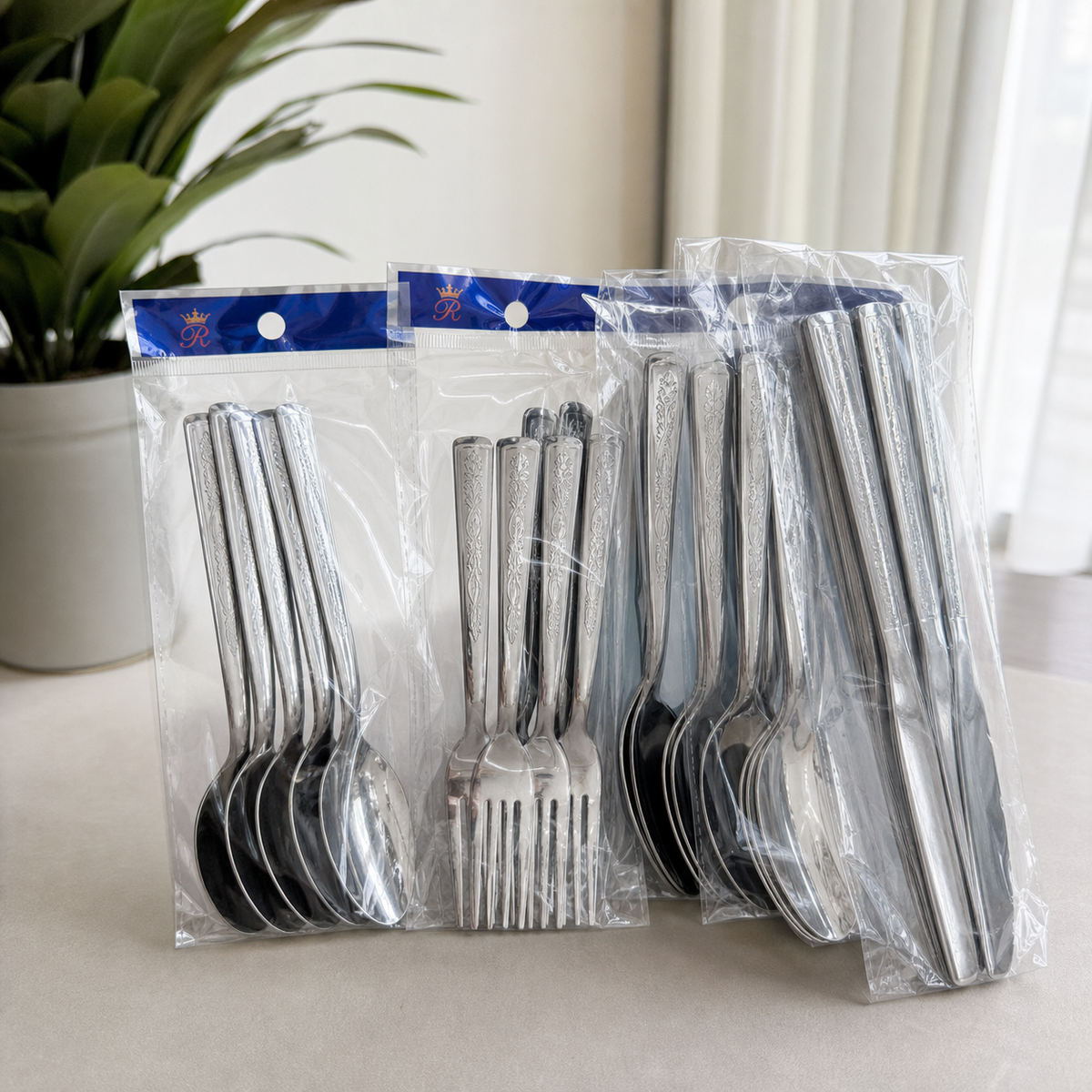Sealed bags of silver-tone cutlery with embossed handles, on a table beside a green plant