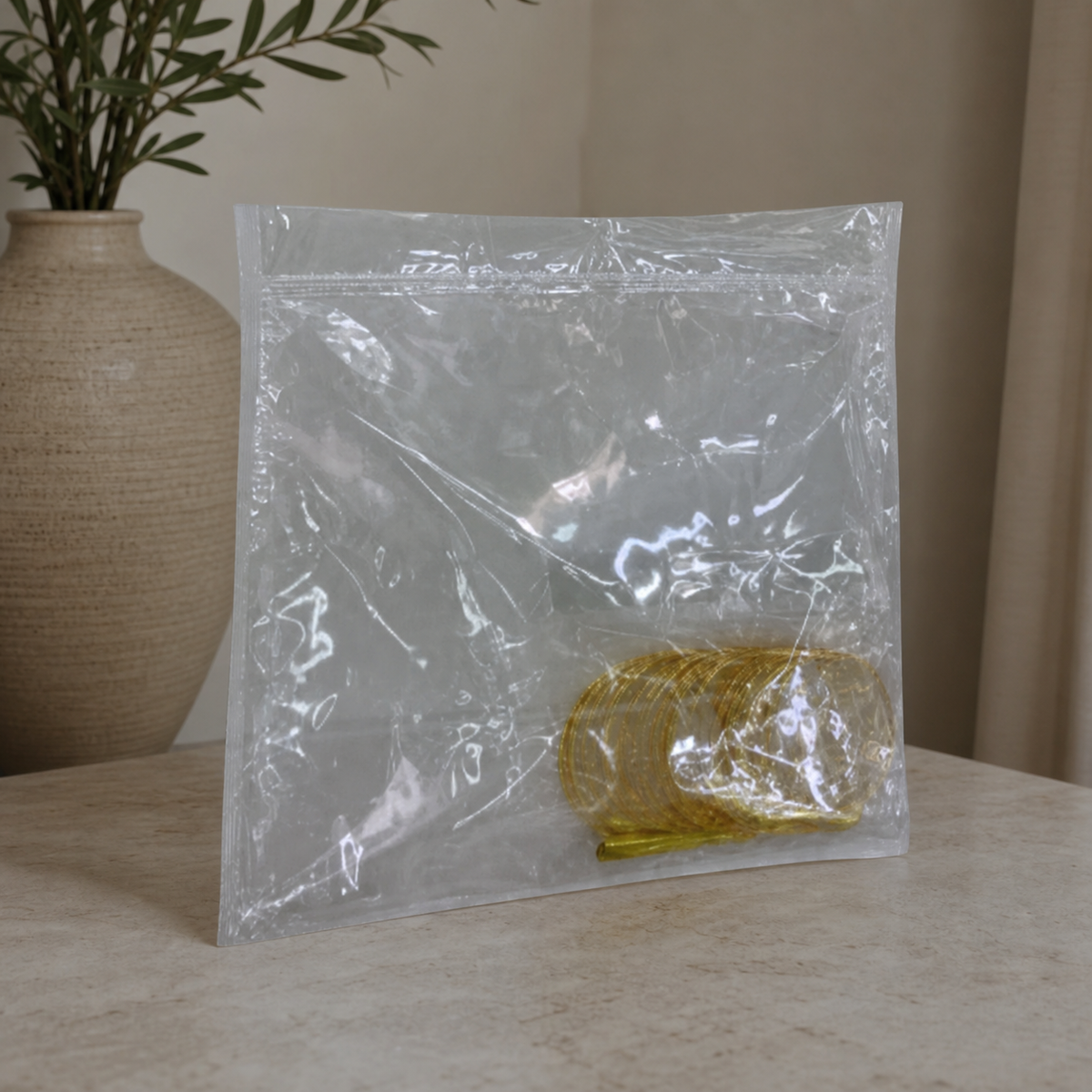 Clear zip-top bag of gold-tone hoops standing on a stone table beside a ribbed vase