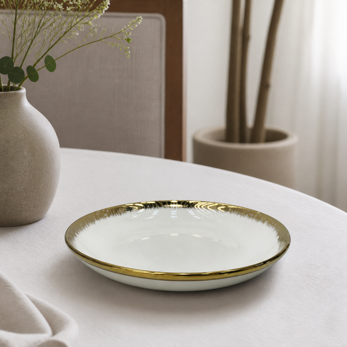 Deep white ceramic coupe plate with brush-stroke gold rim on a dining table beside a stoneware vase