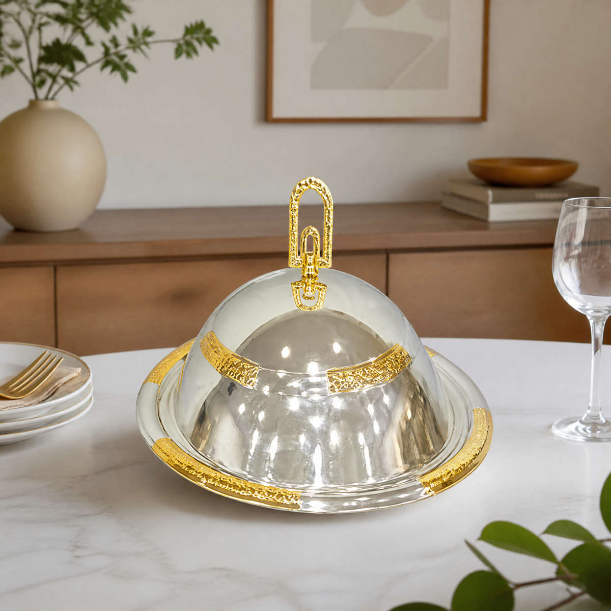 Silver and gold covered food warmer on a set dining table with plates and a wine glass