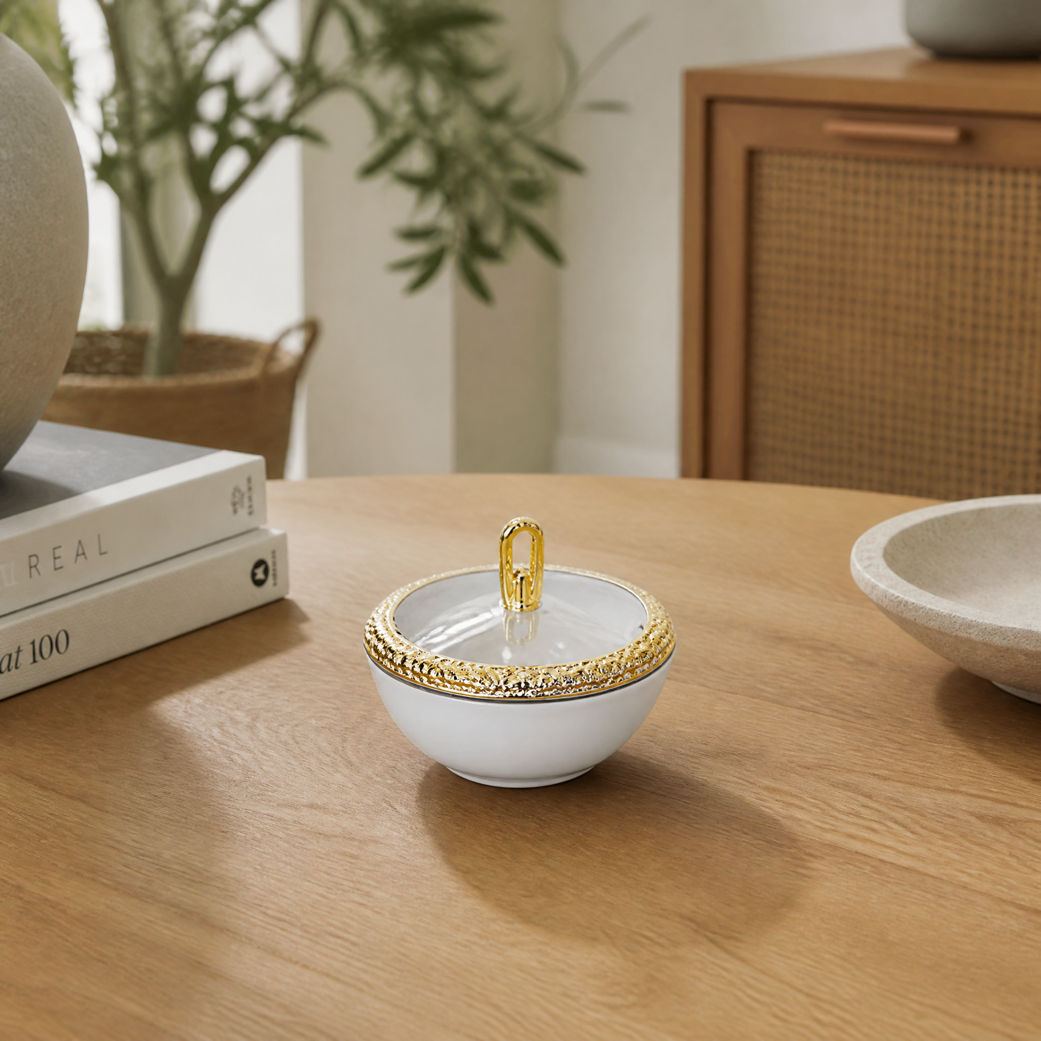 Lidded white and gold bowl on a wooden table beside books and a potted plant