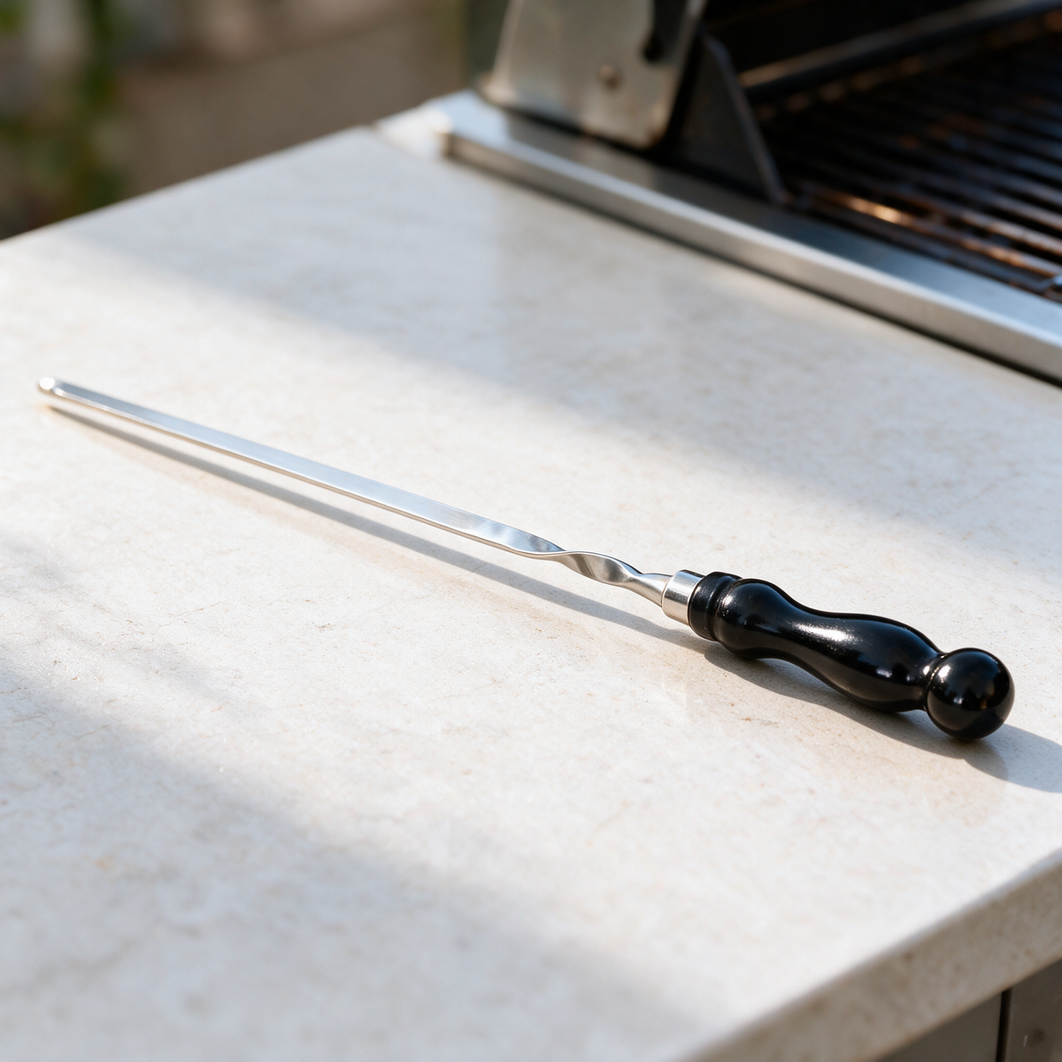 Flat polished skewer with a twisted shaft and plain black handle on a counter beside a grill